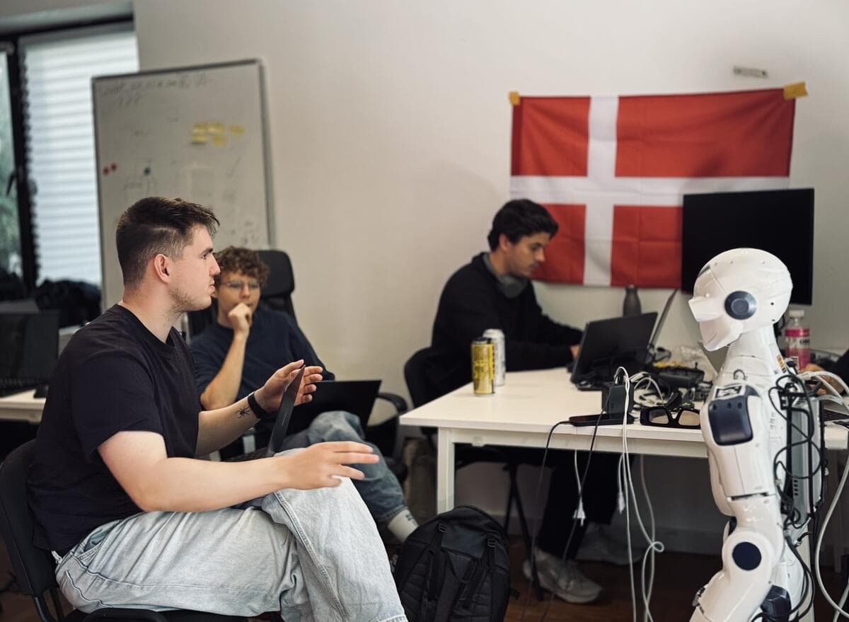 A team at a desk next to a white humanoid robot, with a Danish flag on the wall
